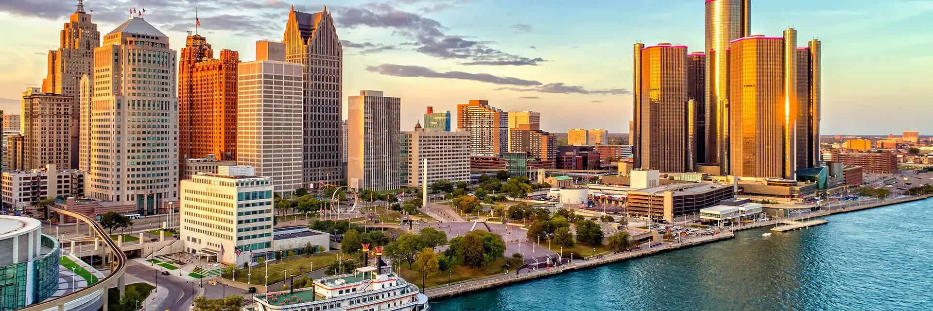 A city skyline with tall buildings and a riverfront at sunset, featuring a boat on the water.