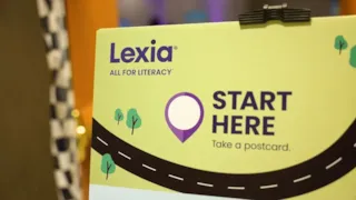 A sign reading Lexia All for Literacy. Start Here. Take a postcard. with a road and trees illustration.