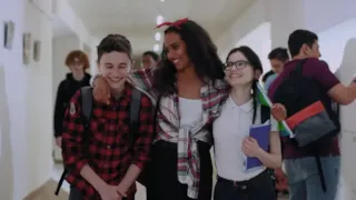 Three smiling teenagers walk down a school hallway, arms around each other, surrounded by other students.