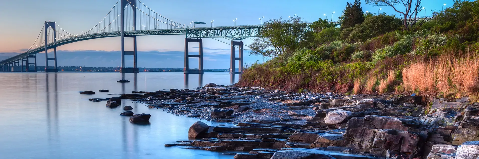 A suspension bridge spans calm water near a rocky shoreline with trees and grass at sunset.