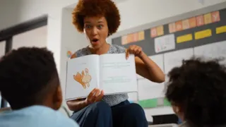A teacher excitedly reads an illustrated book to two young children seated on the floor in a classroom.