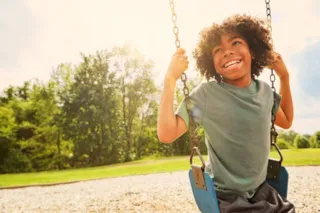 Smiling child with curly hair swings outdoors on a sunny day, surrounded by greenery and a clear sky.