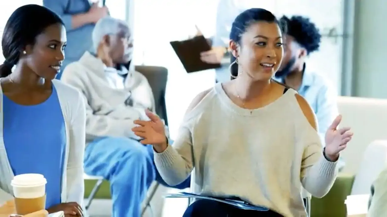 Two women talk and gesture animatedly in a group setting, with other people seated and talking in the background.