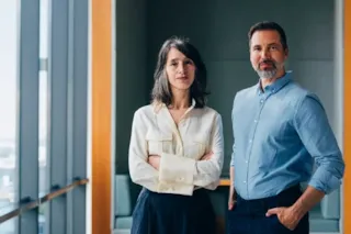 Two professionally dressed adults stand confidently in a modern office with large windows and natural light.