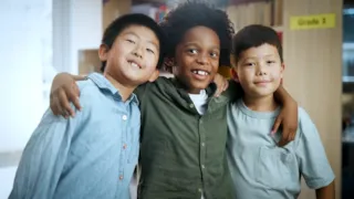 Three young boys smiling with their arms around each other in a classroom setting.