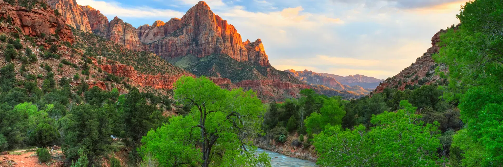 Sunlit red rock mountains and green trees surround a winding river under a blue sky with clouds at sunset.