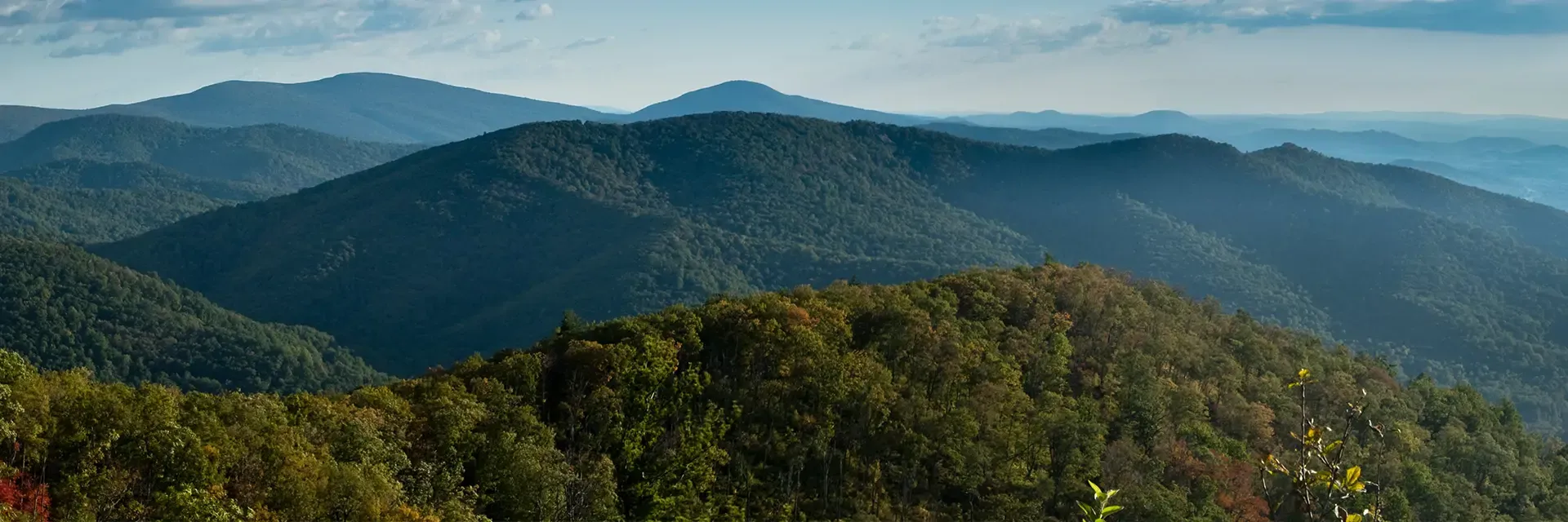 A panoramic view of green, tree-covered mountains under a blue sky with scattered clouds.