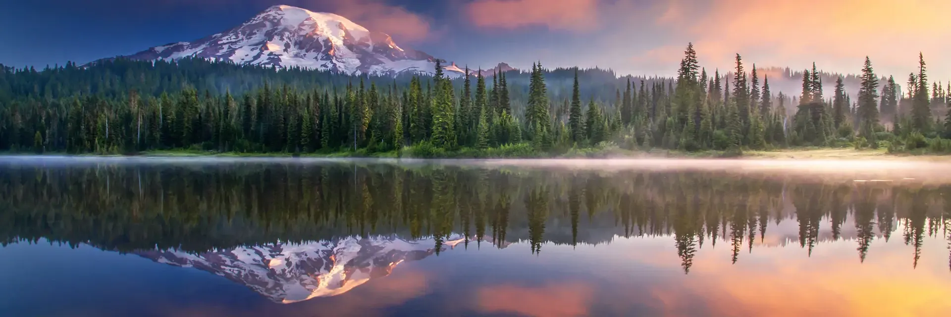 Snow-capped mountain reflected in a calm lake, surrounded by pine trees at sunrise with colorful clouds.