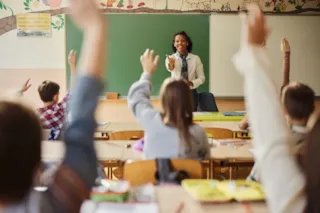 Students in a classroom raise their hands while a teacher stands at the front, smiling and interacting with them.