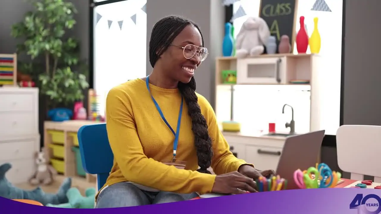 Smiling woman with glasses and braided hair uses a laptop in a colorful classroom decorated with toys and crafts.