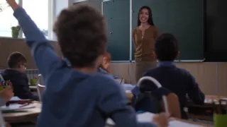 A teacher smiles at the front of a classroom, as a student raises their hand. Students are seated at desks with books and papers.