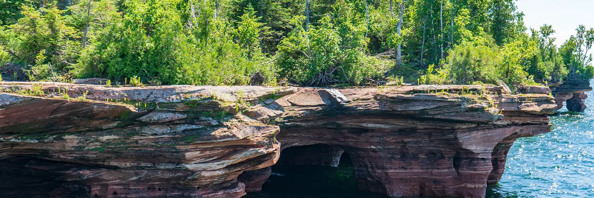 Red sandstone cliffs with a small archway, topped by green trees, beside blue water on a sunny day.