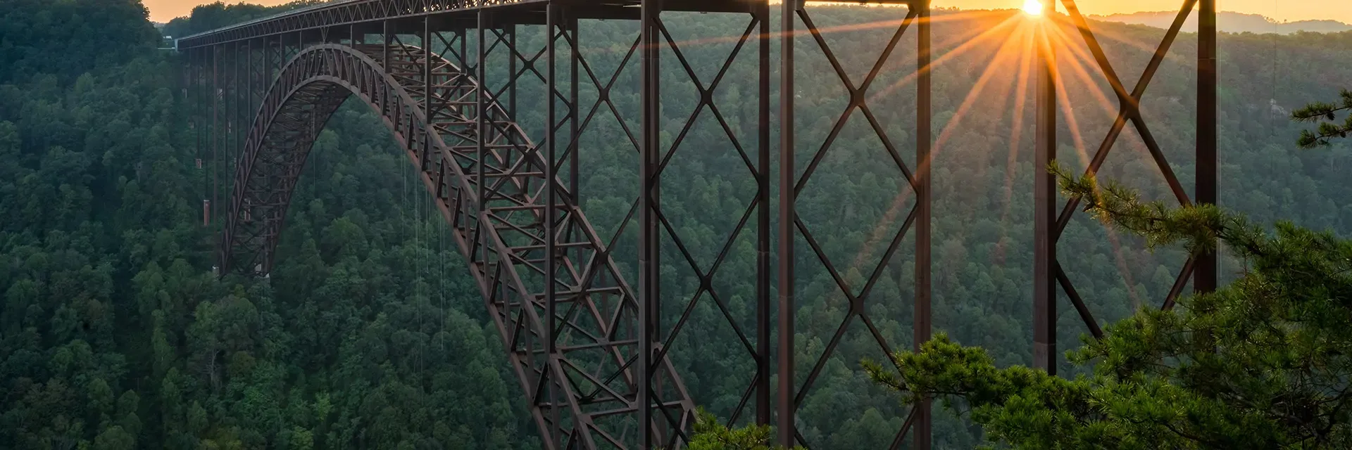 Steel arch bridge spans over a lush forest valley at sunset, with the sun peeking through the bridge’s beams.