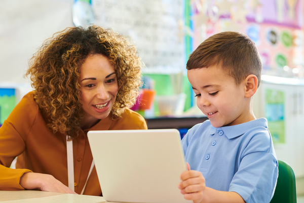 An educator assists a young student with classroom work on a tablet computer