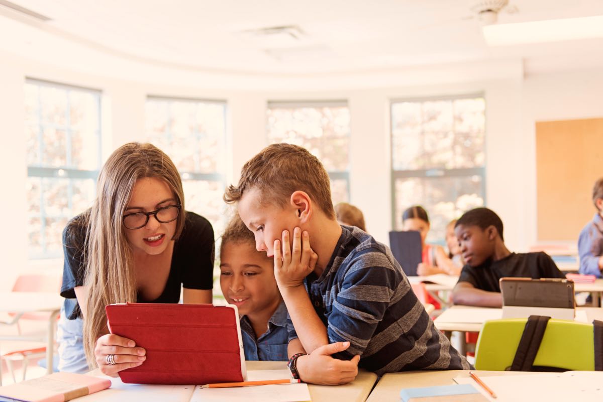 In a classroom, a teacher engages with two grade school students over a tablet.